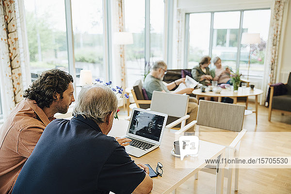Man assisting elderly grandfather using laptop at dining table in retirement home
