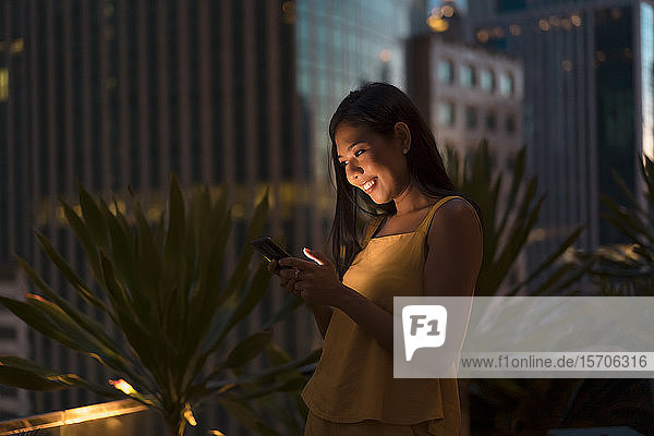 Porträt einer glücklichen Frau  die in der Abenddämmerung auf der Dachterrasse steht und auf ihr Handy schaut  Bangkok  Thailand