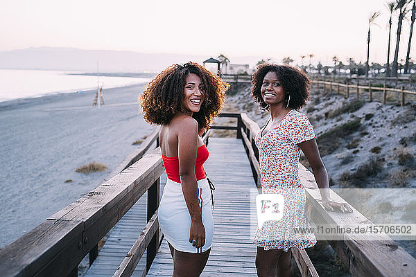 Junge lächelnde Frauen  die abends gemeinsam in Strandnähe gehen  sich umdrehen und in die Kamera schauen
