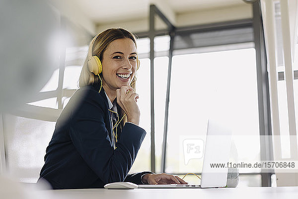 Portrait of happy young businesswoman with headphones and laptop at desk in office