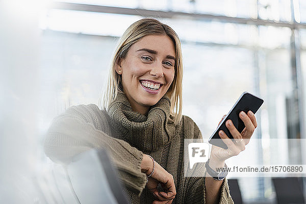 Porträt einer glücklichen jungen Frau  die mit einem Mobiltelefon im Wartebereich sitzt