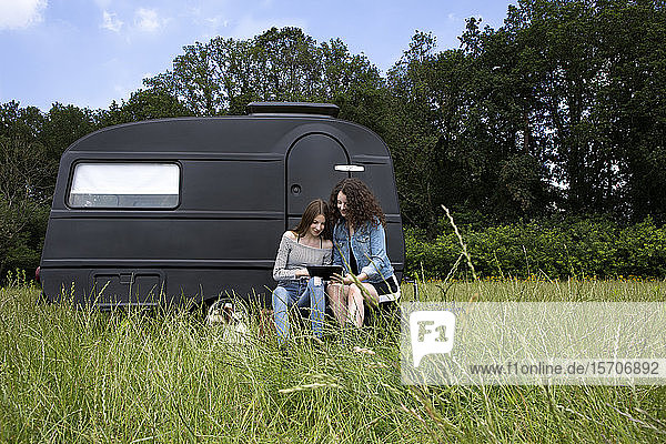 Zwei beste Freunde sitzen auf einer Wiese vor schwarzem Wohnwagen mit digitalem Tablet
