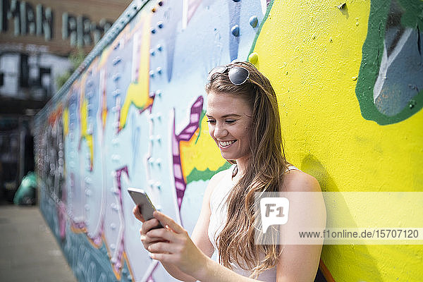 Lächelnde junge Frau benutzt Smartphone an Graffiti-Wand