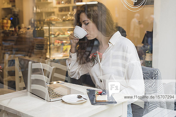 Frau mit Laptop trinkt Kaffee hinter einer Fensterscheibe in einem Cafe