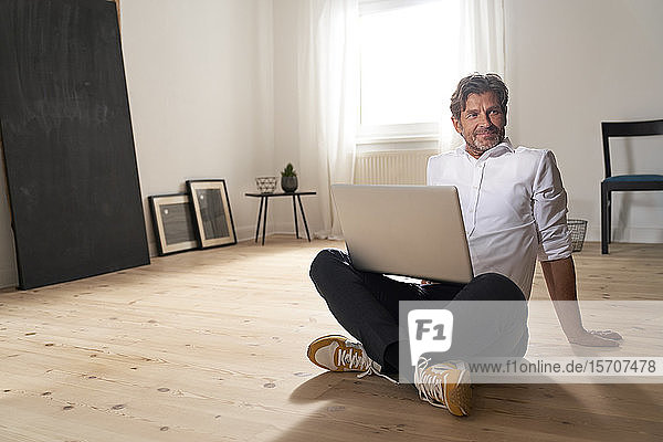 Porträt eines lächelnden reifen Mannes mit Laptop  der zu Hause auf dem Boden sitzt
