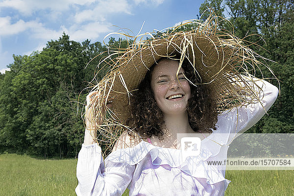 Porträt einer lachenden jungen Frau mit ausgefranstem Strohhut in der Natur