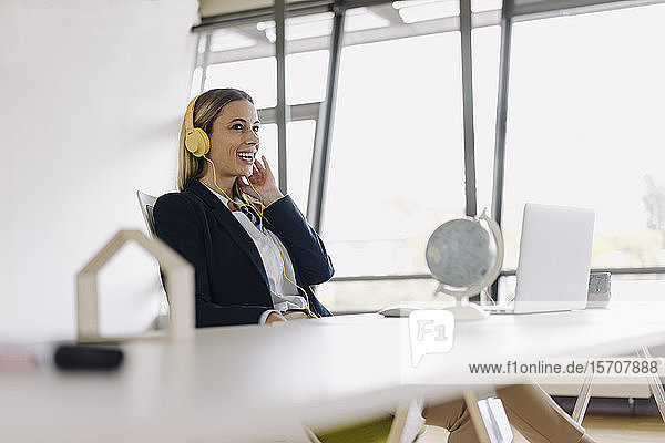 Fröhliche junge Geschäftsfrau mit Kopfhörern und Laptop am Schreibtisch im Büro