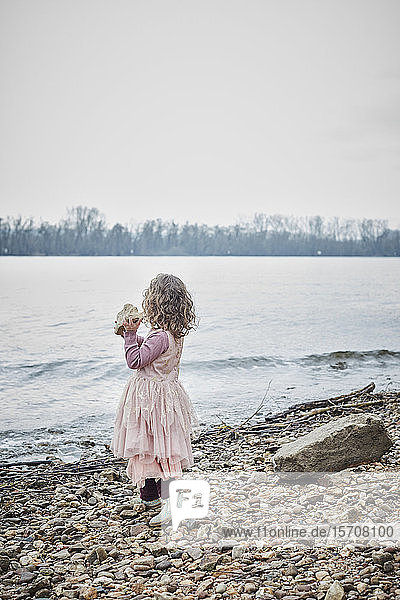 Back view of little girl wearing pink fancy dress costume standing at riverside holding a stone