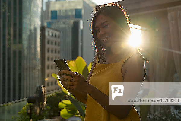 Porträt einer glücklichen Frau auf der Dachterrasse im Gegenlicht mit Blick auf ihr Handy