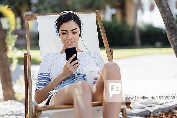 Junge Frau sitzt in einem Liegestuhl  trägt Kopfhörer und benutzt ein Smartphone