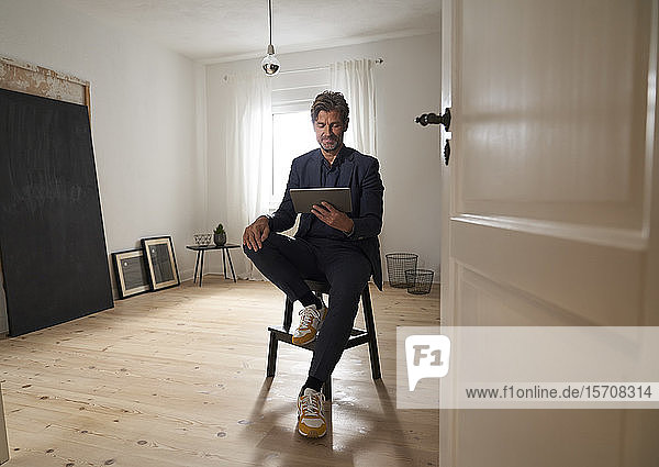 Porträt eines lächelnden reifen Mannes  der zu Hause auf einem Hocker sitzt und auf ein digitales Tablet schaut