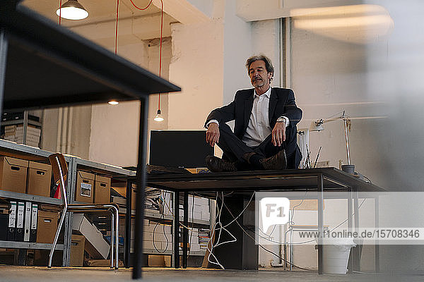 Senior businessman sitting cross-legged on desk in office meditating