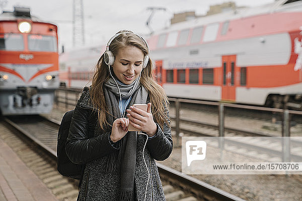 Smiling young woman standing on platform using smartphone and headphones  Vilnius  Lithuania