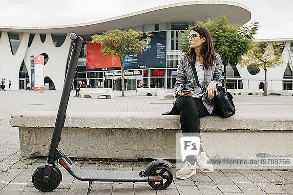 Young businesswoman holding smartphone next to an e-scooter in the city