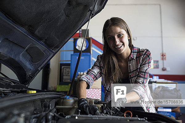 Junge Gelegenheitsfrau lächelt in die Kamera  während sie den Automotor repariert und im Autoservice arbeitet