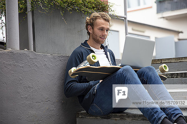 Young man sitting on steps  using laptop on longboard