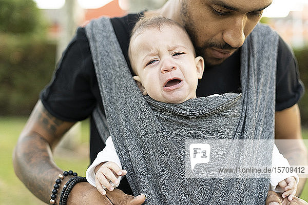 Junger Vater trägt seinen weinenden Sohn in einem Tragetuch
