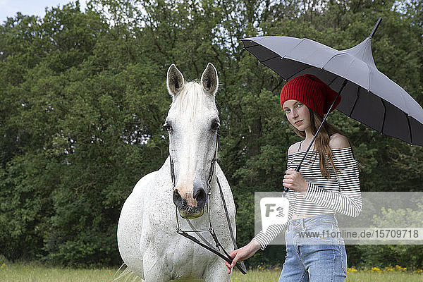 Porträt eines jugendlichen Mädchens auf einer Wiese stehend mit Pferd und Schirm