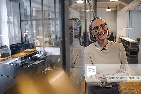 Porträt einer lächelnden reifen Geschäftsfrau  die sich im Büro an eine Glasscheibe lehnt
