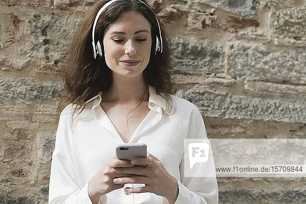 Young woman with smartphone and headphones outdoors at a stone wall