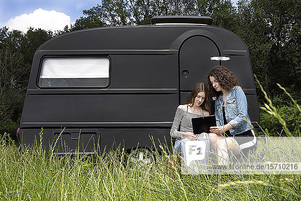 Zwei Freunde sitzen auf einer Wiese vor einem schwarzen Wohnwagen und benutzen ein digitales Tablett