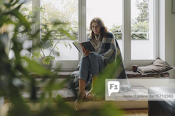 Eine reife Frau sitzt auf einer wunderbaren Fensterbank  in eine Decke gewickelt  liest ein Buch