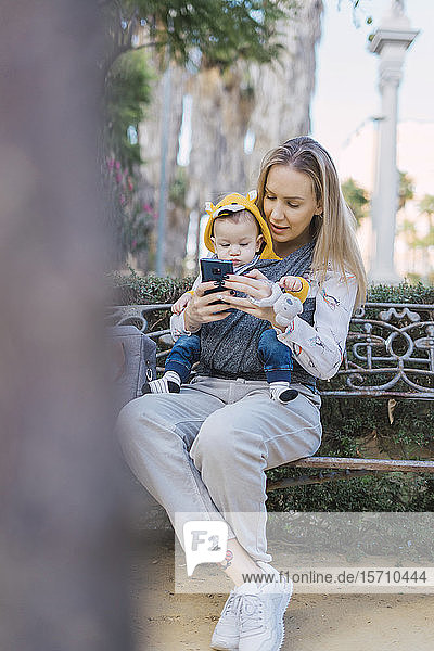 Mutter ruht sich mit dem kleinen Jungen auf einer Parkbank mit Hilfe eines Smartphones aus