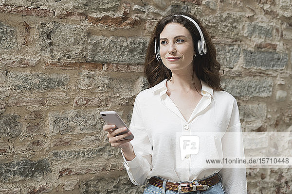 Young woman with smartphone and headphones outdoors at a stone wall