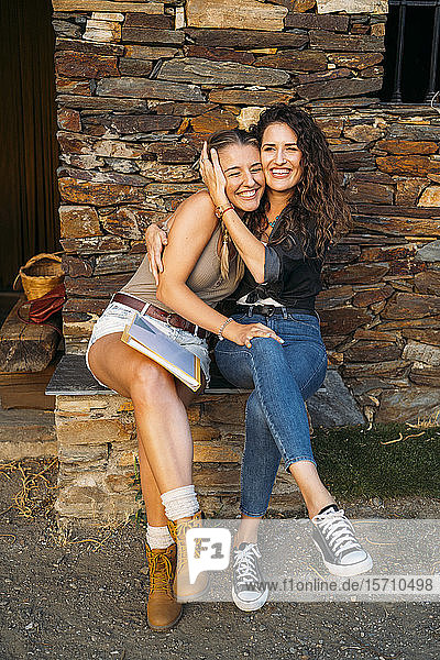 Zwei fröhliche Frauen mit einer Mappe umarmen sich an einem Steinhaus