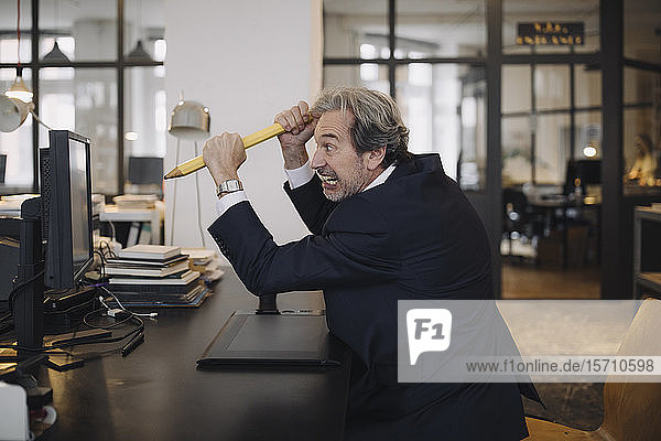 Angry senior businessman with giant pencil at desk in office