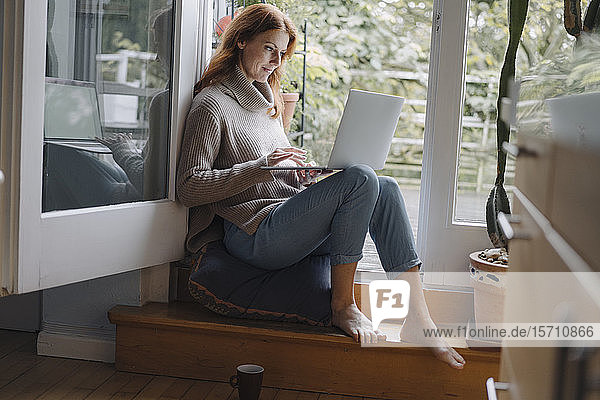 Eine reife Frau sitzt auf den Stufen einer Balkontür und benutzt einen Laptop