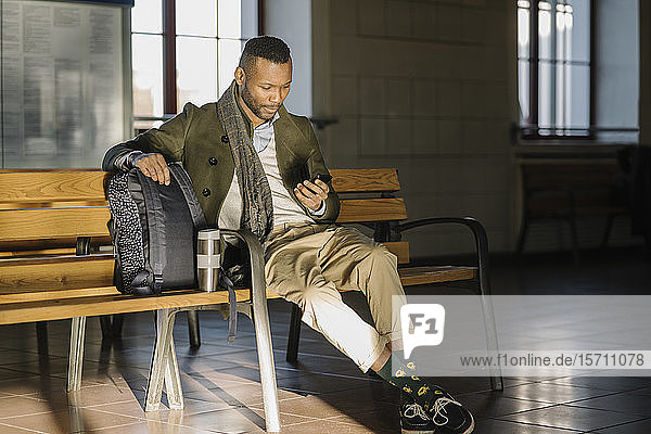 Stylischer Mann benutzt Smartphone  während er auf einer Bank in einem Bahnhof sitzt
