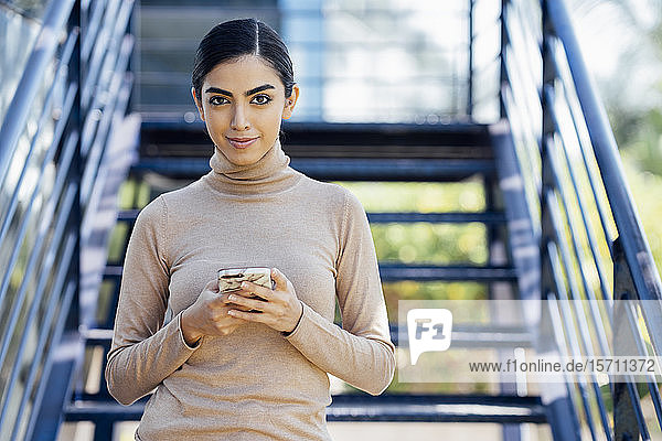 Portrait of confident young woman holding smartphone on exterior stairs