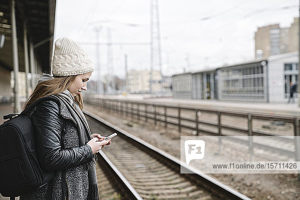 Lächelnde junge Frau mit Rucksack steht mit dem Handy auf dem Bahnsteig