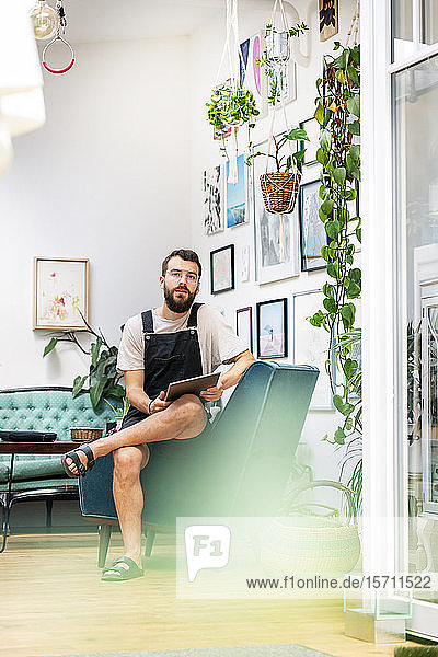 Junger Mann in Latzhose sitzt in seinem Wohnzimmer und benutzt ein digitales Tablett
