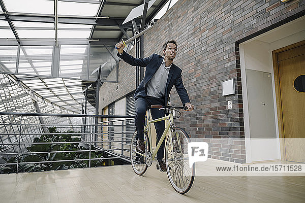 Geschäftsmann hält ein Spielzeugschwert und fährt Fahrrad in modernem Bürogebäude