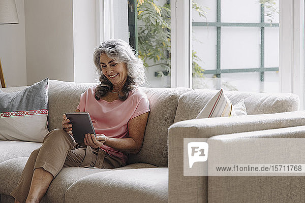 Smiling mature woman using tablet sitting on couch at home