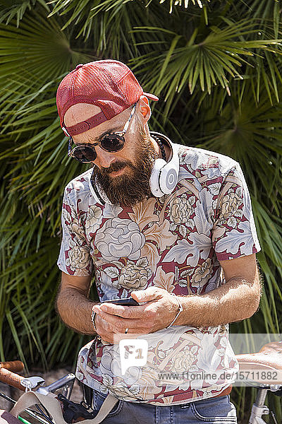 Reifer Mann mit roter Basecap  Sonnenbrille und weißen Kopfhörern mit Smartphone