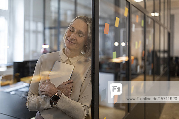 Mature businesswoman with closed eyes leaning against glass pane in office