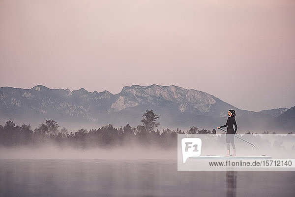 Frau steht auf und paddelt auf dem Kirchsee im Morgennebel  Bad Tölz  Bayern  Deutschland