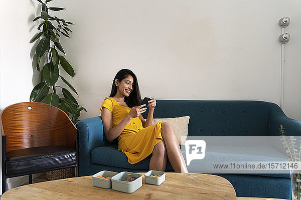 Lächelnde junge Frau sitzt auf Couch und benutzt Mobiltelefon