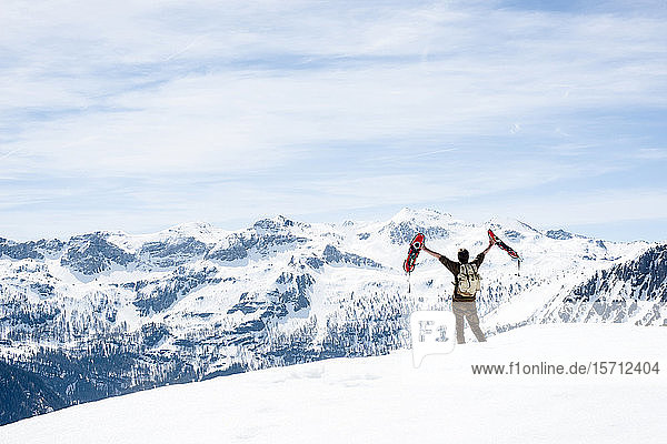 Mann mit Rucksack,  auf Berggipfel stehend,  winkt mit seinen Schneeschuhen,  Rückansicht,  Bundesland Salzburg,  Österreich