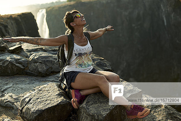 Glückliche Frau sitzt mit offenen Armen auf einem Felsen an den Victoria-Fällen bei Sonnenuntergang  Simbabwe