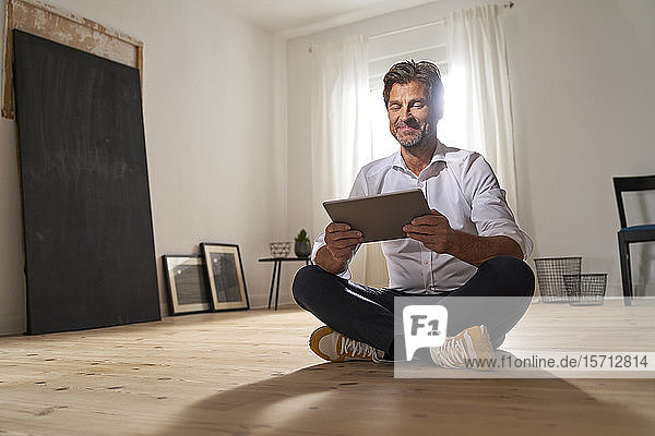 Porträt eines lächelnden  reifen Mannes  der zu Hause auf dem Boden sitzt und ein digitales Tablet benutzt