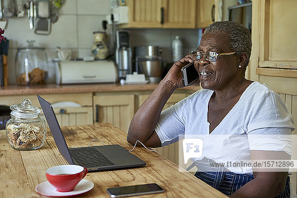 Ältere Frau sitzt am Küchentisch mit Laptop und Smartphone