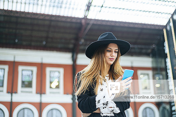 Junge Frau trägt einen Hut und benutzt Mobiltelefon am Bahnhof