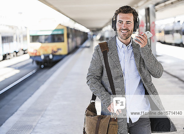 Lächelnder junger Geschäftsmann mit Handy und Kopfhörern am Bahnhof