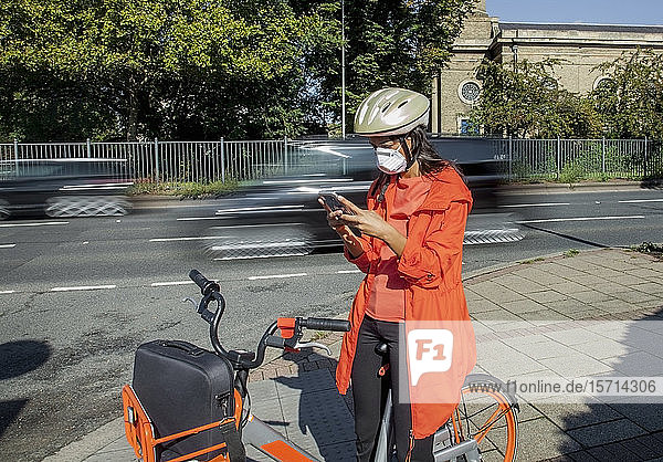 Young woman with helmet and face mask  sitting on bicycle  using smartphone