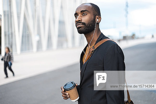 Portrait of young businessman with coffee to go listening music with earphones outdoors