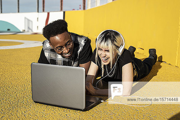Lachendes junges Paar  das mit Kopfhörer und Laptop im Freien Spaß hat